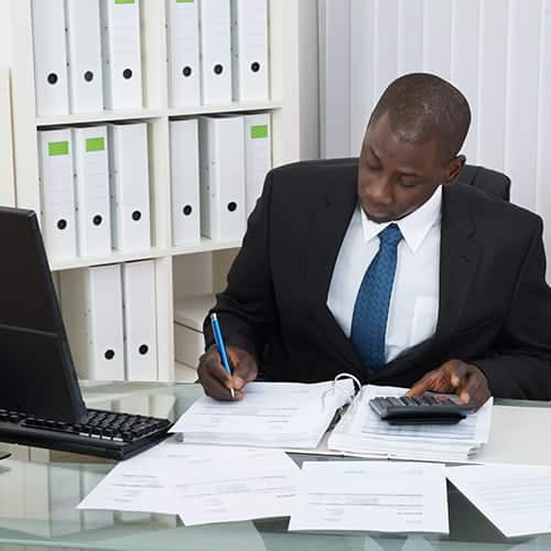 A businessman with a calculator calculating finances on documents with a keyboard and monitor on his table