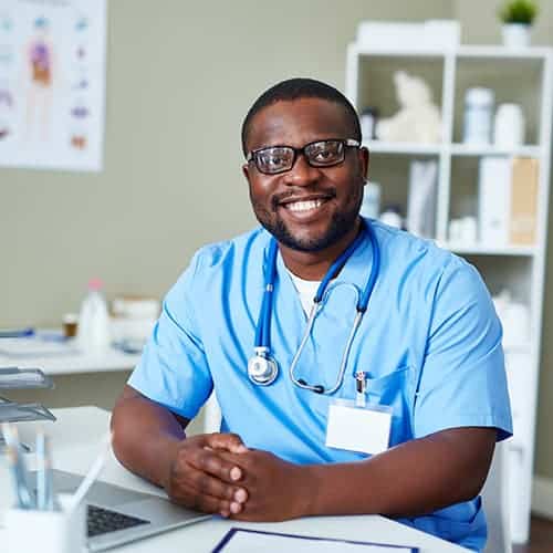 A cheerful male doctor in blue attire with a stethoscope in his neck sitting at his desk and facing the camera